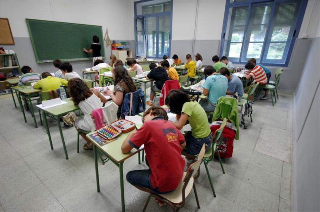 Schulklasse im Klassenzimmer arbeitet konzentriert, währenddessen die Lehrerin etwas an die Tafel schreibt.